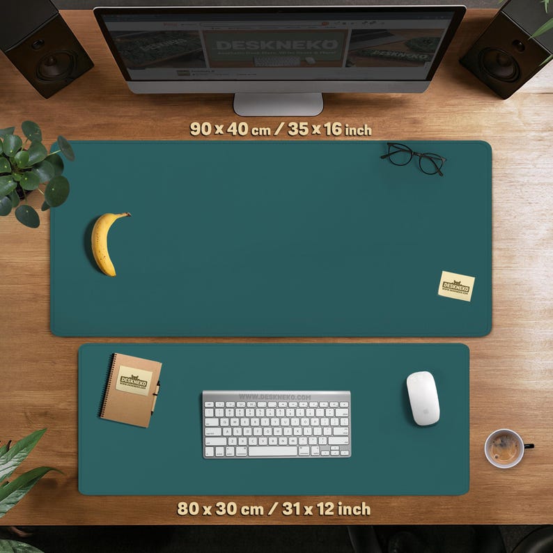 Op de afbeelding: Twee teal bureaumatten, 90 x 40 cm en 80 x 30 cm, worden op een houten bureau getoond. De grotere mat heeft een banaan, een bril en een plakbriefje. De kleinere mat heeft een toetsenbord, muis en notitieboek. De website DESKNEKO is zichtbaar.