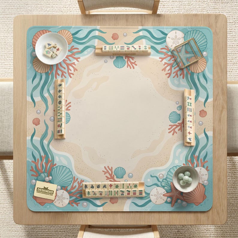 May include: A square mahjong mat featuring a sandy beach center surrounded by a border of teal waves, coral, seashells, and sand dollars. The mat includes four wooden tile racks, two white bowls with mahjong tiles and green glass beads, and a small card.