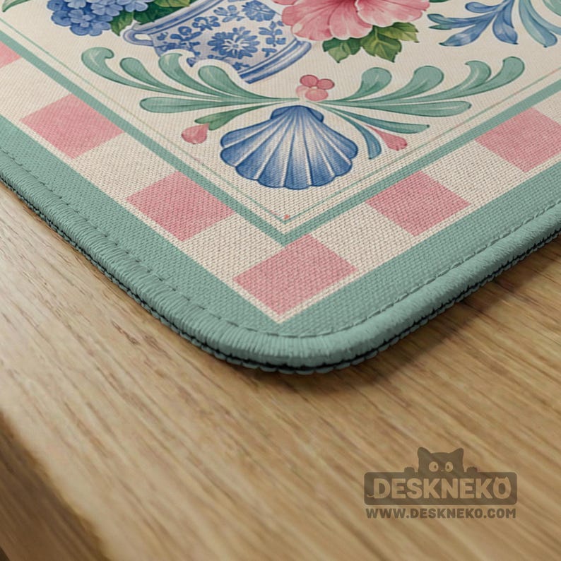 May include: A decorative desk mat featuring a floral design with pink and blue flowers, a blue seashell, and a pink and white checkered border. The mat has a light green trim and the Deskneko logo.