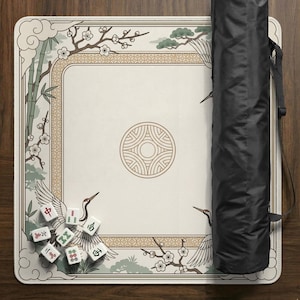 May include: A cream-colored Mahjong mat with an Asian-inspired design featuring cranes, bamboo, and cherry blossoms. The mat has a decorative border and a central circular emblem. A black carrying case is on the right, and Mahjong tiles are scattered on the mat.