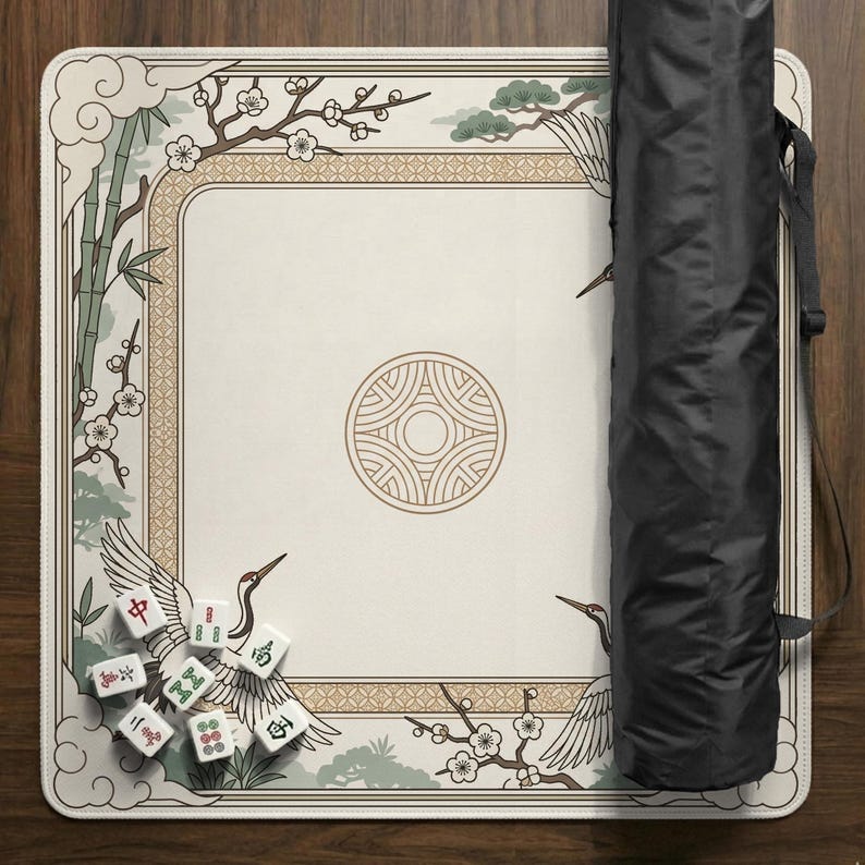 May include: A cream-colored Mahjong mat with an Asian-inspired design featuring cranes, bamboo, and cherry blossoms. The mat has a decorative border and a central circular emblem. A black carrying case is on the right, and Mahjong tiles are scattered on the mat.