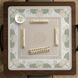 May include: A square mahjong game table with a wooden frame and a beige playing surface. The surface features a floral design with lotus flowers and a central circular pattern. Several mahjong tiles and a small bowl are visible.