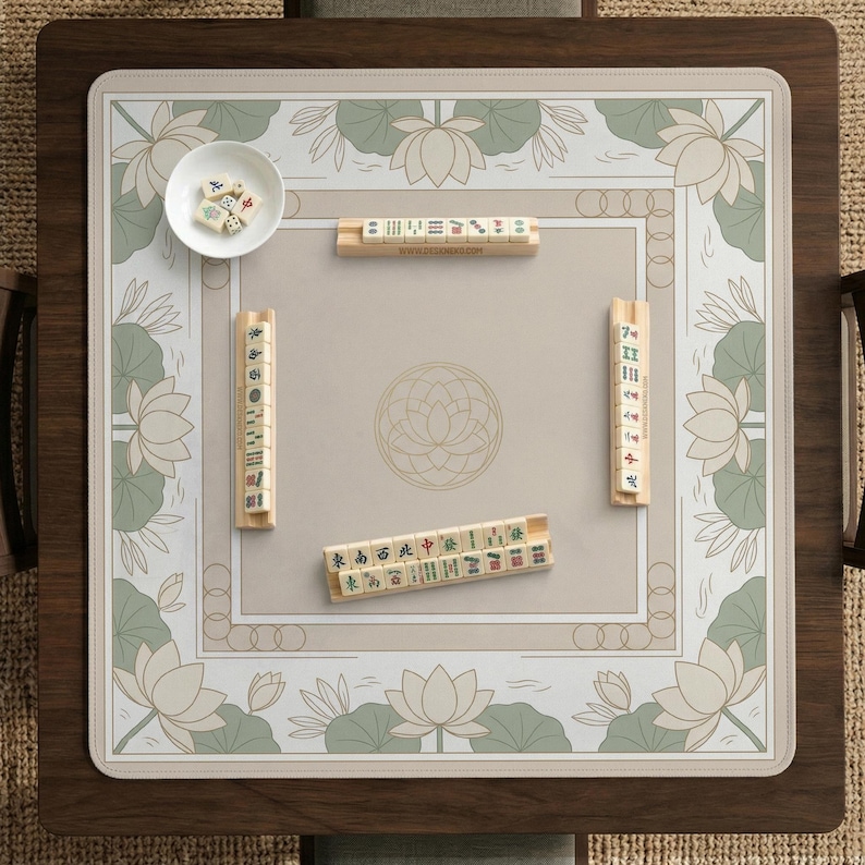 May include: A square mahjong game table with a wooden frame and a beige playing surface. The surface features a floral design with lotus flowers and a central circular pattern. Several mahjong tiles and a small bowl are visible.
