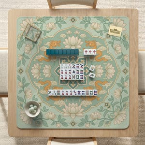 May include: A square mahjong game mat with a floral and dragon design in shades of green, gold, and white. The mat features a set of mahjong tiles, including those with Chinese characters. A small bowl with white spheres is in the lower left corner.