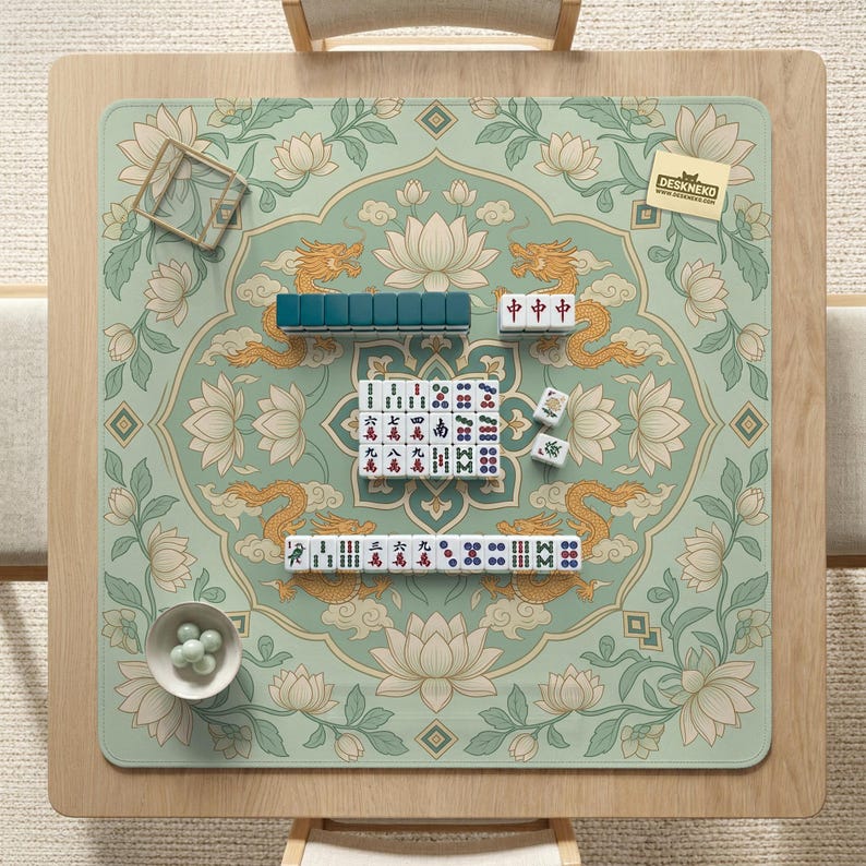 May include: A square mahjong game mat with a floral and dragon design in shades of green, gold, and white. The mat features a set of mahjong tiles, including those with Chinese characters. A small bowl with white spheres is in the lower left corner.