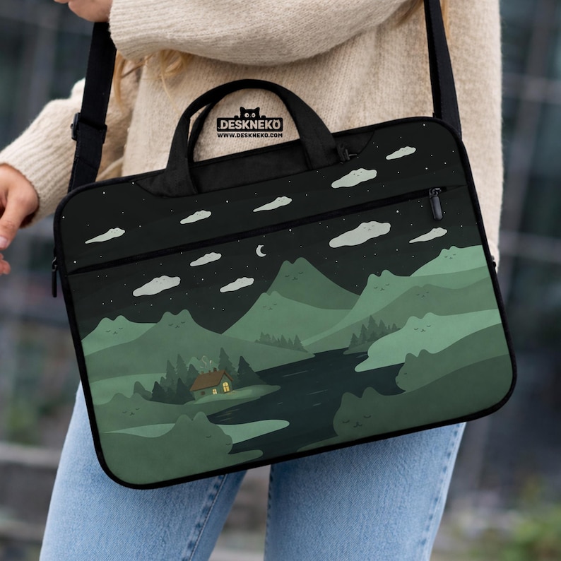 May include: Black laptop bag with a scenic landscape design. The design features a night sky with clouds, stars, and a crescent moon over green mountains, a river, and a cabin with a lit window. The bag has a handle and a shoulder strap. The brand name "DESKNEKO" is printed on the top.