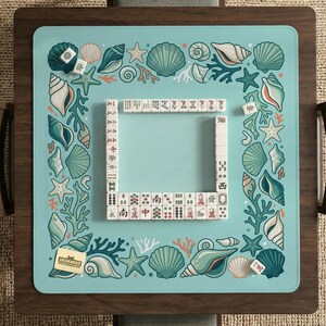May include: A square Mahjong game mat in light blue, decorated with seashell and coral designs. The mat has a central playing area framed by a border of illustrations. Mahjong tiles are arranged on the mat, ready for play.