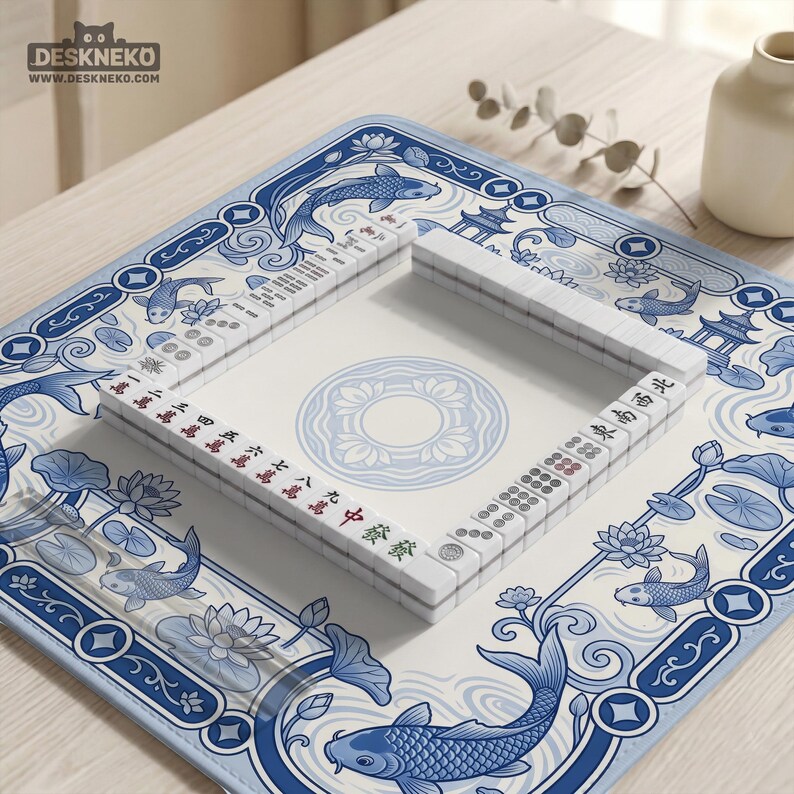 May include: A square mahjong mat featuring a blue and white pattern of koi fish, lotus flowers, and pagodas. The mat displays a central circular lotus motif and is surrounded by a border of swimming fish. White mahjong tiles are arranged in a square.