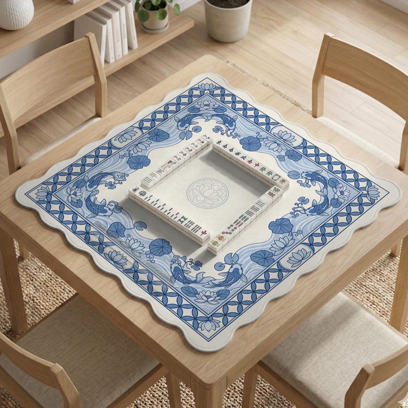 May include: A square wooden table with a blue and white patterned mat featuring koi fish and lotus flowers. A Mahjong set is placed on the mat. The table is surrounded by wooden chairs.
