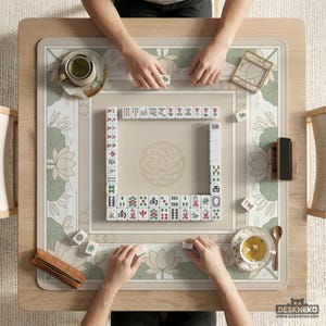 May include: Overhead view of a Mahjong game in progress. The game is set on a light wood table with a beige and green floral patterned mat. Players are arranging tiles, with cups of tea and accessories nearby. The game is in play.