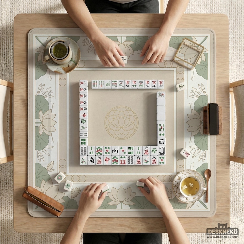 May include: Overhead view of a Mahjong game in progress. The game is set on a light wood table with a beige and green floral patterned mat. Players are arranging tiles, with cups of tea and accessories nearby. The game is in play.