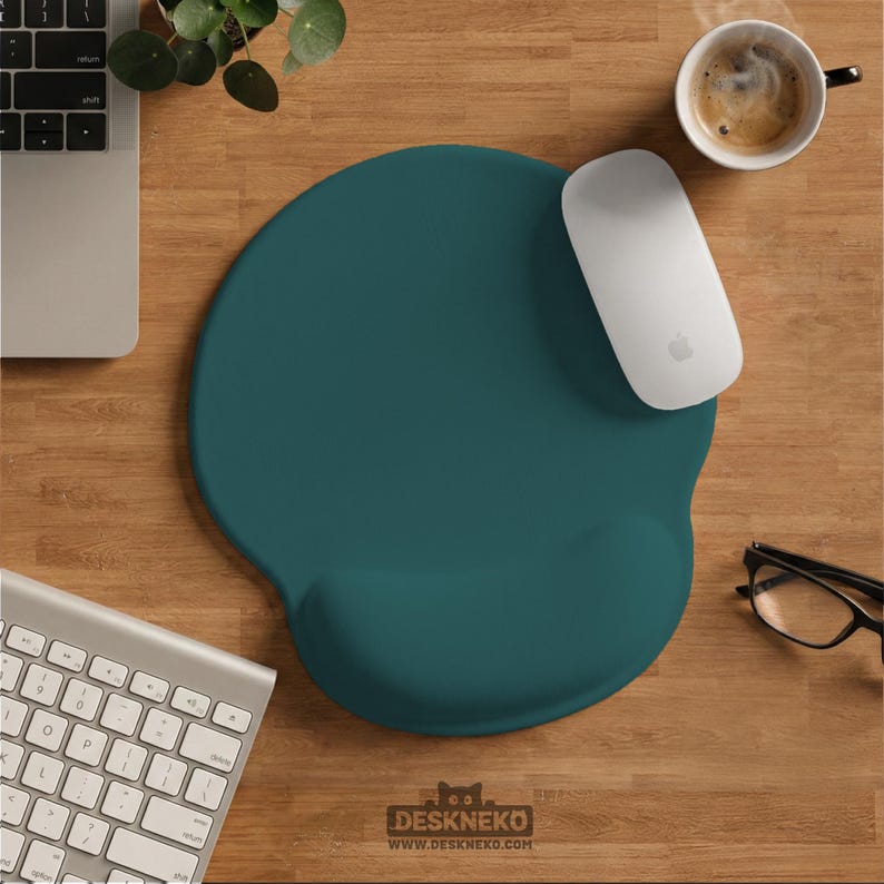 May include: Teal ergonomic mouse pad with a wrist rest, a white Apple mouse, and a keyboard on a wooden desk. A cup of coffee and glasses are also on the desk. The mouse pad has the text "DESKNEKO" and the website address.
