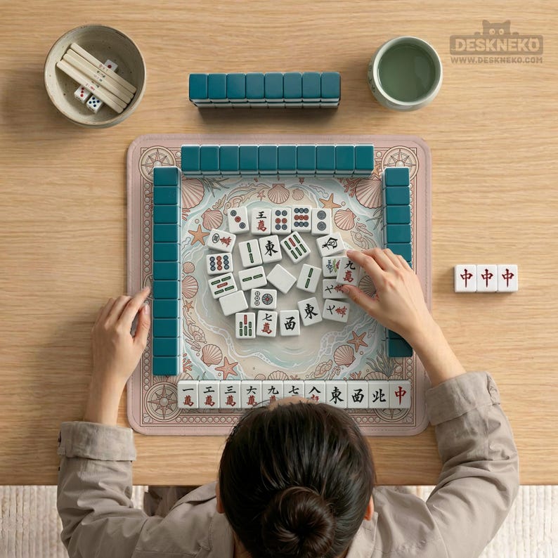 May include: A top-down view of a Mahjong set on a wooden table. The set includes teal tiles, a pink mat with a seashell design, dice, and a small bowl. A person is arranging the tiles. A cup of tea is also visible.