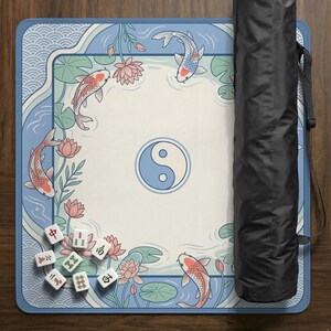 May include: A rectangular Mahjong mat with a serene design featuring koi carp, lotus flowers, and a yin-yang symbol. The mat is bordered in blue with a wave pattern. A black carrying case is rolled up next to the mat. Mahjong tiles are scattered on the mat.
