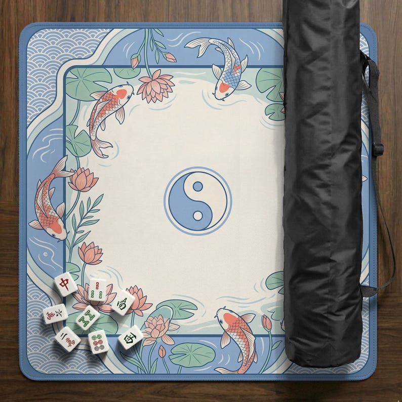 May include: A rectangular Mahjong mat with a serene design featuring koi carp, lotus flowers, and a yin-yang symbol. The mat is bordered in blue with a wave pattern. A black carrying case is rolled up next to the mat. Mahjong tiles are scattered on the mat.