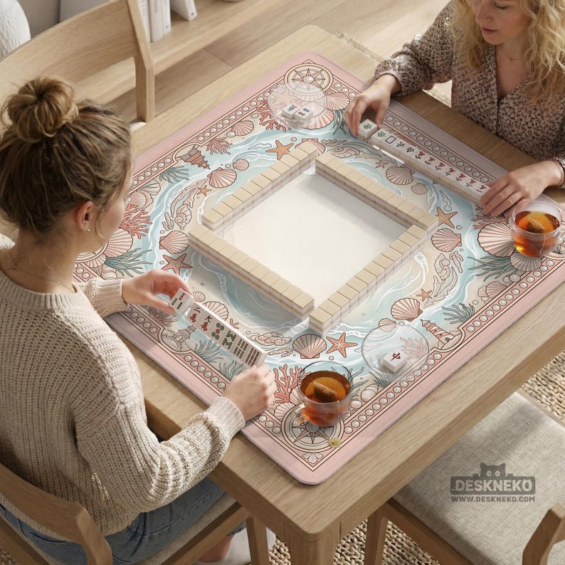 May include: A Mahjong game in progress on a wooden table. The game mat features a sea-themed design with seashells, starfish, and waves in pastel colors. The game includes wooden tiles and tea cups.