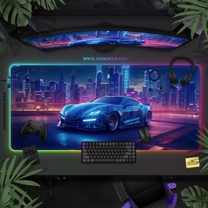 May include: A large rectangular gaming mousepad featuring a futuristic car design with a city skyline backdrop. The mousepad has a neon-colored border and includes a keyboard, mouse, controller, and headset. The website address www.deskneko.com is visible.