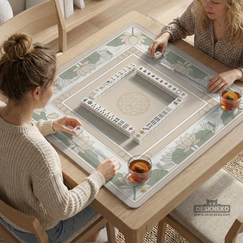 May include: A beige mahjong set with white tiles is displayed on a wooden table. The table has a beige mat with a lotus flower design. Two people are playing mahjong. The website address deskneko.com is visible.