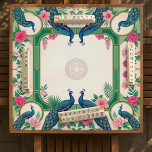 May include: A square mahjong table featuring a cream-colored surface with a decorative border of blue peacocks, green foliage, and pink hibiscus flowers. Four wooden tile racks are positioned on the edges, each holding a row of ivory-colored mahjong tiles.