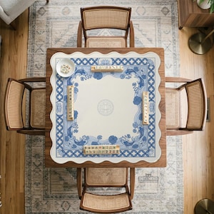 May include: Overhead view of a square wooden table set for a game. The table has a white and blue patterned mat with a floral and koi fish design. Four cane-backed chairs surround the table. A small bowl and game pieces are on the mat.