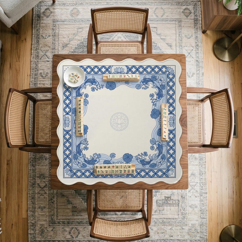 May include: Overhead view of a square wooden table set for a game. The table has a white and blue patterned mat with a floral and koi fish design. Four cane-backed chairs surround the table. A small bowl and game pieces are on the mat.