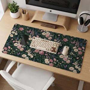Op de afbeelding: Een bureauonderlegger met bloemenpatroon, met een donkergroene achtergrond en roze, witte en paarse bloemen. Een wit toetsenbord, muis en smartphone liggen op de mat. De mat ligt op een houten bureau.