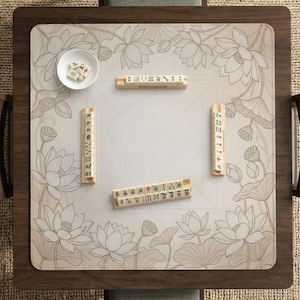 Puede incluir: Una mesa de Mahjong de madera con una esterilla beige con estampado floral con flores de loto. La mesa tiene un marco de madera marr&oacute;n oscuro y contiene varios juegos de fichas de Mahjong y un cuenco blanco peque&ntilde;o con dados.