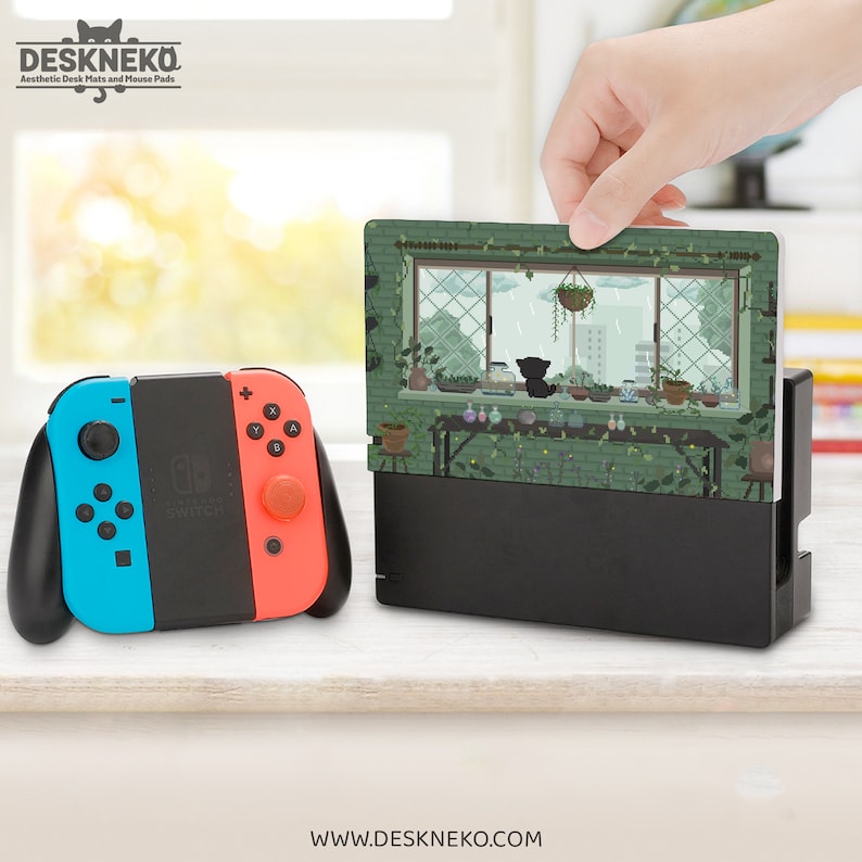 May include: A Nintendo Switch console with blue and red controllers is next to a black charging dock. A decorative card featuring a cat looking out a window is inserted into the dock. The text "DESKNEKO" and "www.DESKNEKO.com" are visible.