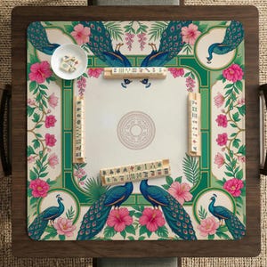 May include: A square mahjong mat featuring a cream-colored center with a circular mandala design, surrounded by a green border decorated with blue peacocks and pink hibiscus flowers. Four wooden tile racks hold mahjong tiles on the mat.
