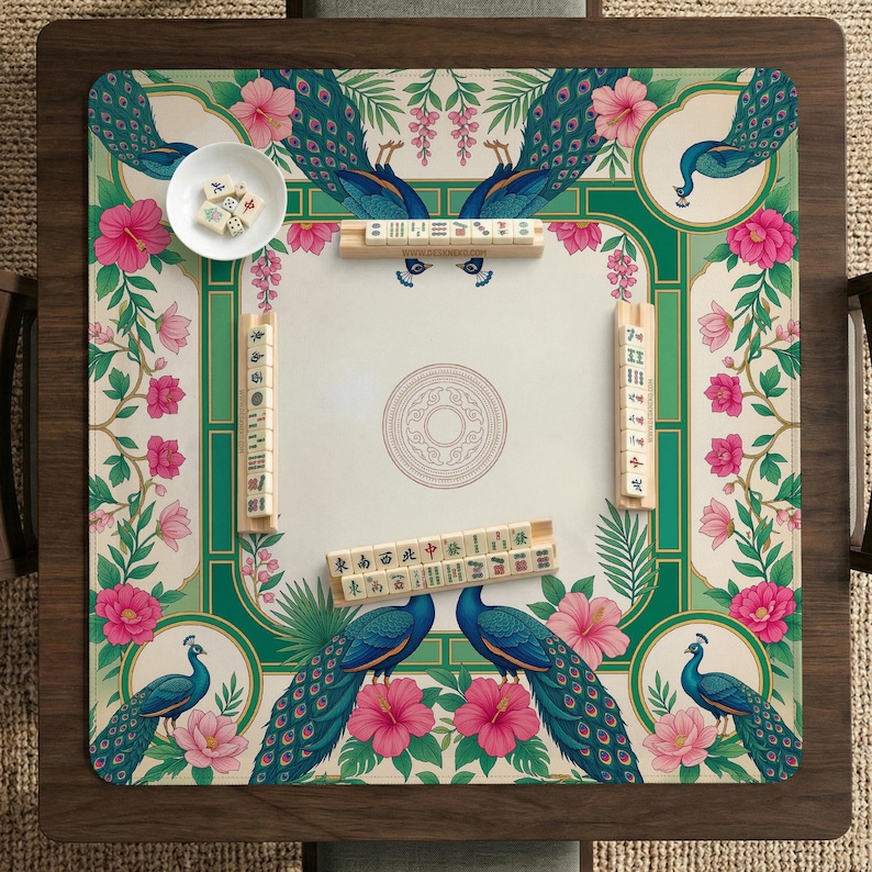 May include: A square mahjong mat featuring a cream-colored center with a circular mandala design, surrounded by a green border decorated with blue peacocks and pink hibiscus flowers. Four wooden tile racks hold mahjong tiles on the mat.