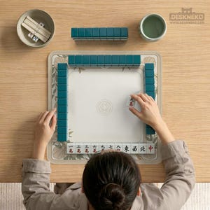 May include: A top-down view of a Mahjong set on a wooden table. The set includes teal tiles, dice, and a floral-patterned mat. A person is arranging a tile. A small bowl and a cup are also present.