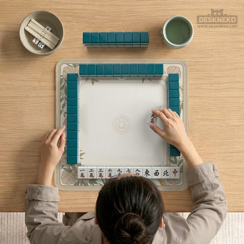 May include: A top-down view of a Mahjong set on a wooden table. The set includes teal tiles, dice, and a floral-patterned mat. A person is arranging a tile. A small bowl and a cup are also present.