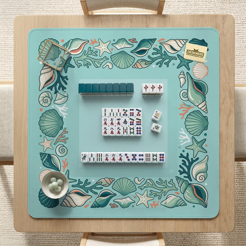 May include: A light blue mahjong mat with a seashell and coral border. The mat features a set of mahjong tiles and accessories. The mat is on a wooden table. The brand name "DESKNEKO" is visible.