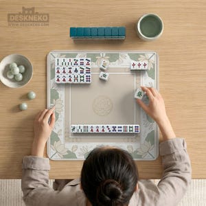 May include: A person playing Mahjong on a beige mat with a floral border. The game includes white tiles with red, green, and blue markings, arranged on the mat. A teal tile holder and a small green bowl are also visible.