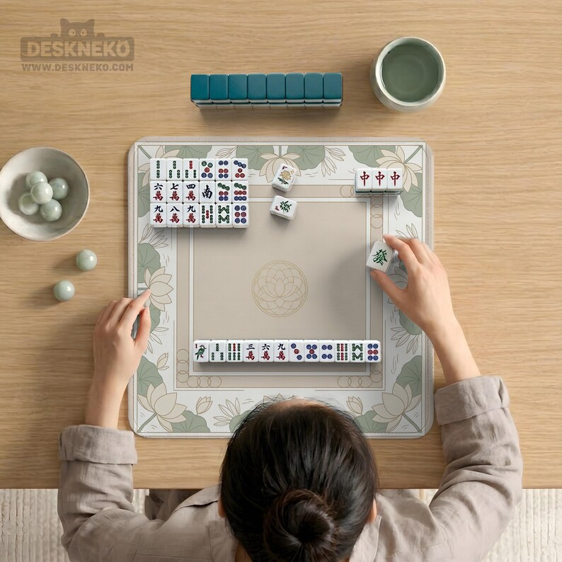 May include: A person playing Mahjong on a beige mat with a floral border. The game includes white tiles with red, green, and blue markings, arranged on the mat. A teal tile holder and a small green bowl are also visible.
