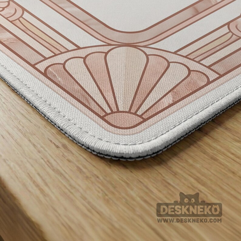 May include: A close-up of a rectangular desk mat with a geometric art deco design in shades of peach and white. The mat has a stitched black border. The Deskneko logo and website are visible in the bottom right corner.