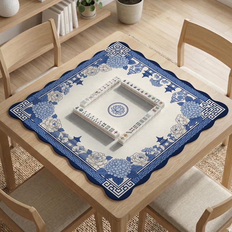 May include: A square mahjong table mat with a blue and white floral design. The mat features a scalloped edge and a central lotus flower motif. Mahjong tiles are arranged on the mat, ready for play.