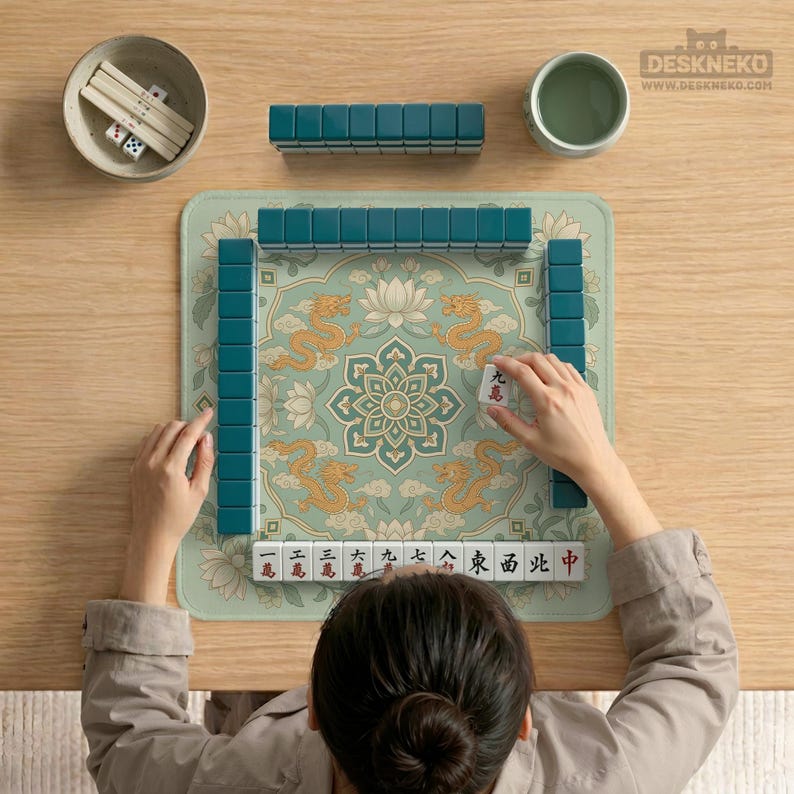 May include: A mahjong set featuring a square play mat with a light green background, a central lotus flower, and golden dragon illustrations. The set includes teal tiles, a bowl with white dice, and a small cup, all arranged on a wooden surface.