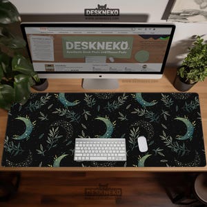 Op de afbeelding: Een zwarte bureaumat met een hemels ontwerp met halve manen, sterren en bladeren. Een wit toetsenbord en muis liggen op de mat. De mat ligt op een houten bureau met een computermonitor en planten.