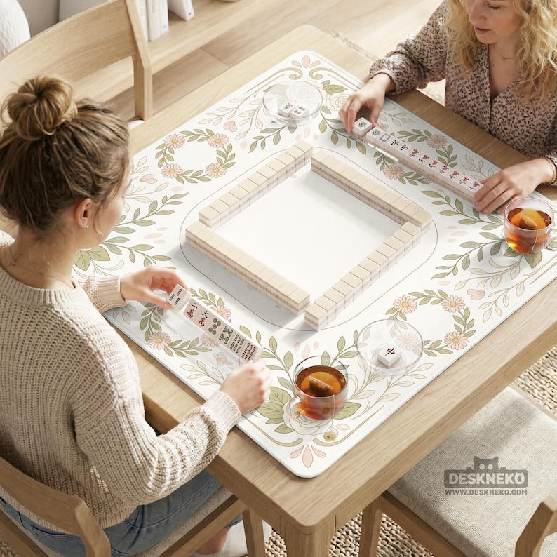 May include: A square mahjong mat featuring a white surface with a floral and leaf pattern in soft green and peach tones. The mat displays a central arrangement of rectangular mahjong tiles, with additional tiles held by players and tea cups.