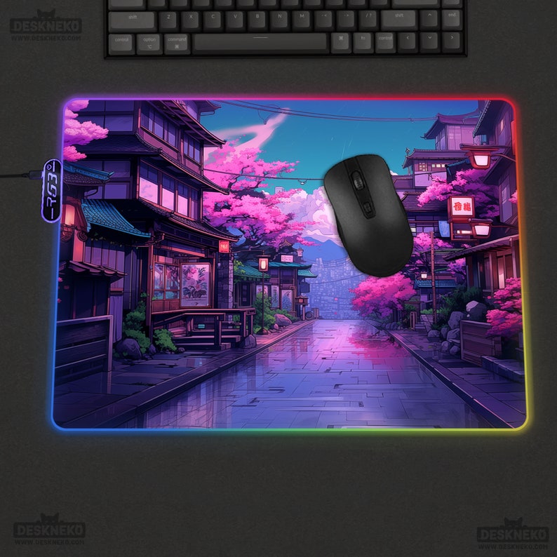 May include: A rectangular gaming mouse mat featuring a colourful anime-style illustration of a Japanese street scene with cherry blossom trees. The mat has a LED edge and a black mouse. The text "DESKNEKO" is visible.