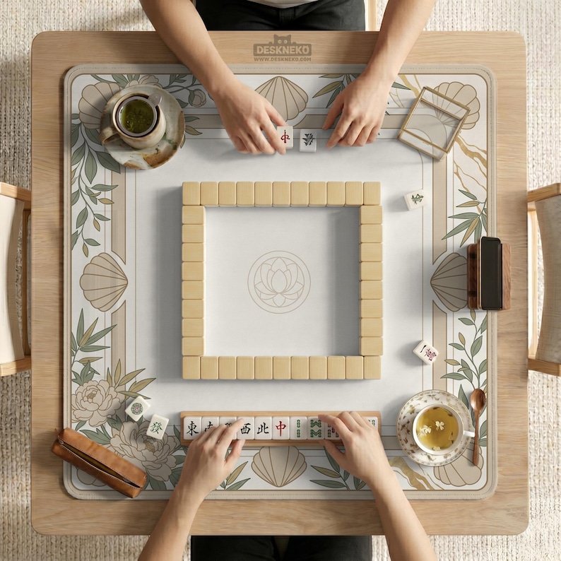 May include: Overhead view of a mahjong table with beige tiles and a floral and seashell patterned mat. The table is made of light wood and has the text "DESKNEKO" on the top. Two people are playing, with hands visible, and tea cups are on the table.