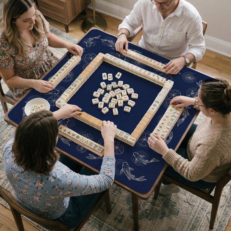 Puede incluir: Cuatro personas jugando al Mahjong alrededor de una mesa azul oscuro con un dise&ntilde;o de peces koi y flores de loto. Las piezas del juego, beige y blancas, est&aacute;n dispuestas sobre la mesa. Vista superior.