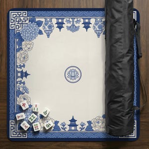 May include: A blue and white Mahjong game mat with floral and pagoda designs. The mat has a cream-colored center with a lotus flower emblem. A black carrying case is visible on the right side. Several Mahjong tiles are scattered on the mat.