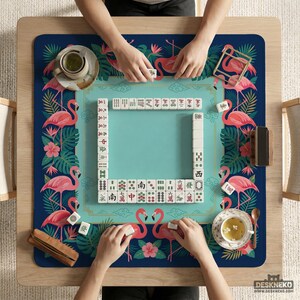 May include: A square mahjong table with a teal playing surface and a navy blue border decorated with pink flamingos and tropical foliage. Players are arranging tiles. The table has a wooden frame and is set with tea cups.