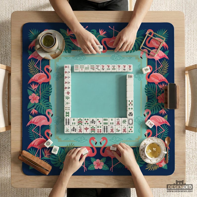 May include: A square mahjong table with a teal playing surface and a navy blue border decorated with pink flamingos and tropical foliage. Players are arranging tiles. The table has a wooden frame and is set with tea cups.