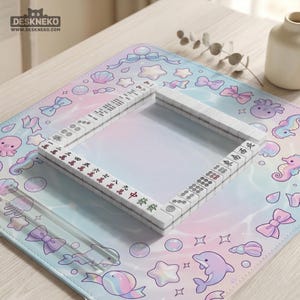 May include: A white Mahjong set with red and black markings sits on a pastel-colored mat. The mat features a repeating pattern of bows, sea creatures, and stars. The set is surrounded by a clear plastic border. The Deskneko logo is in the top left corner.