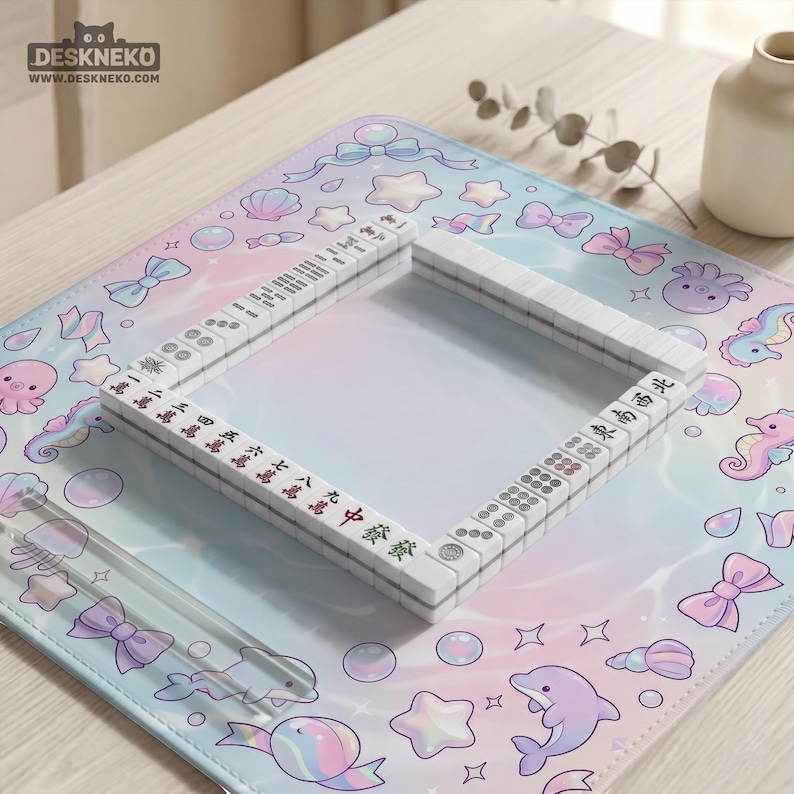 May include: A white Mahjong set with red and black markings sits on a pastel-colored mat. The mat features a repeating pattern of bows, sea creatures, and stars. The set is surrounded by a clear plastic border. The Deskneko logo is in the top left corner.