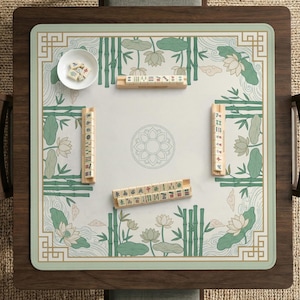 Puede incluir: Una mesa de juego de Mahjong cuadrada con un dise&ntilde;o floral verde y blanco, bamb&uacute; y flores de loto. La mesa tiene un marco de madera e incluye fichas y un cuenco peque&ntilde;o con dados. El juego est&aacute; listo para jugar.