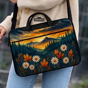 May include: A black laptop bag with a colorful landscape design. The design features a sunset over a mountain range, with trees, flowers, and fall leaves. The bag has a handle and a shoulder strap, and the text "DESKNEKO" is visible.